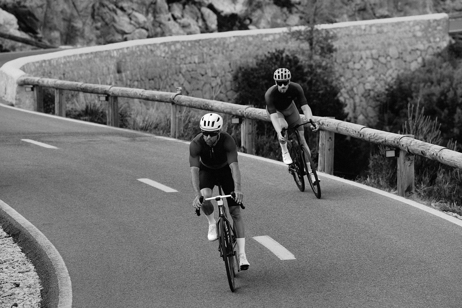 Two cyclists riding on a winding road with a stone wall and vegetation in the background.